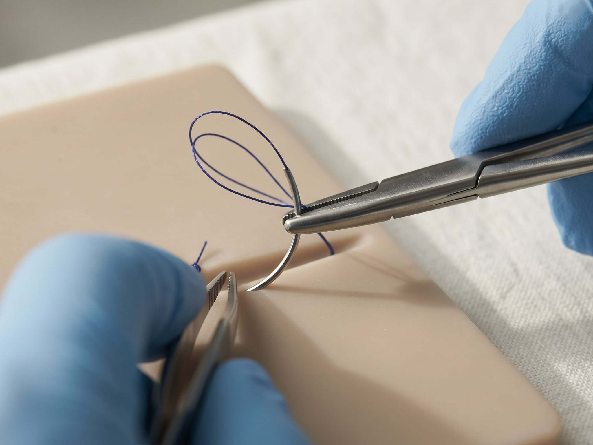 Gloved hands tying a surgical knot with a needle holder on a silicone suture practice pad.